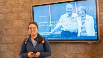 A smiling woman stands with a phone in front of a digital display featuring two people.