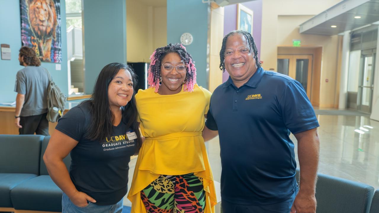 Three smiling adults in a bright lobby; center woman in yellow dress and patterned skirt (photo)