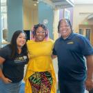 Three smiling adults in a bright lobby; center woman in yellow dress and patterned skirt (photo)